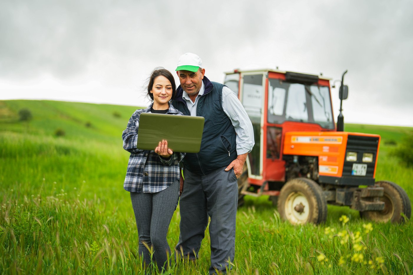 young female agronomist and senior farmer controlling wheat field with laptop with smart farming apps tractor in background